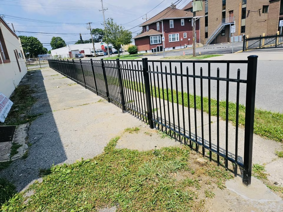 Black metal fence along a sidewalk, with buildings and a street in the background. Green grass and weeds are growing.