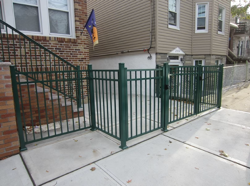 Green metal fence enclosing a small concrete area in front of a house, next to a sidewalk and steps.