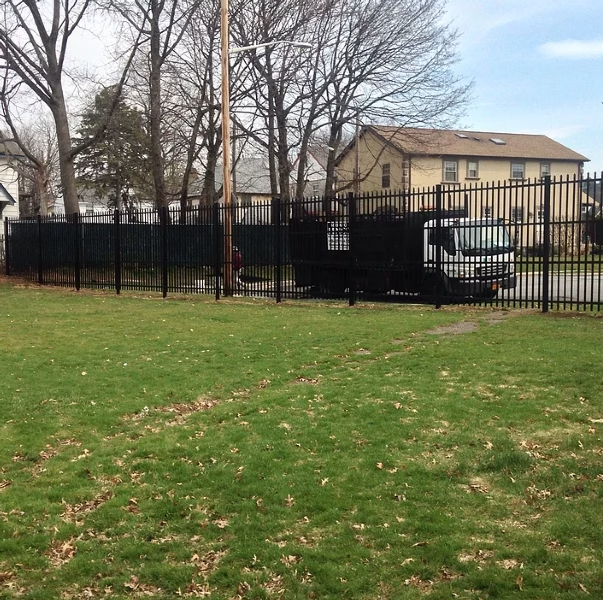 Black metal fence surrounds a grassy area, with a building and trees in the background under a cloudy sky.