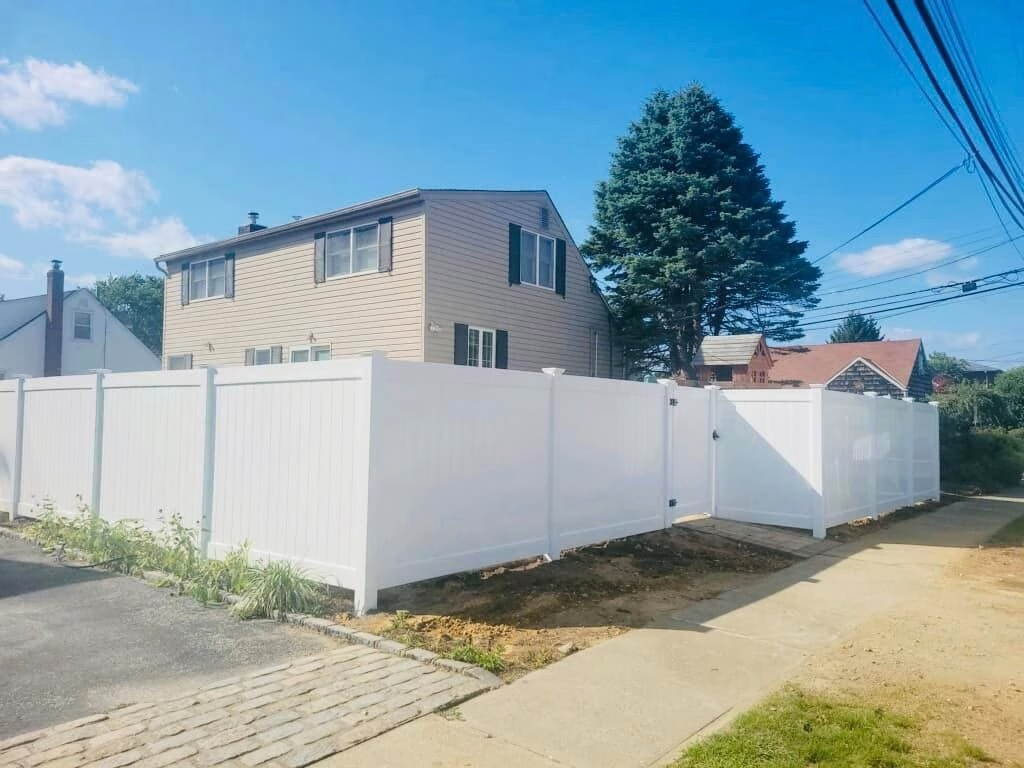 White privacy fence surrounding a house with a green tree on a sunny day.