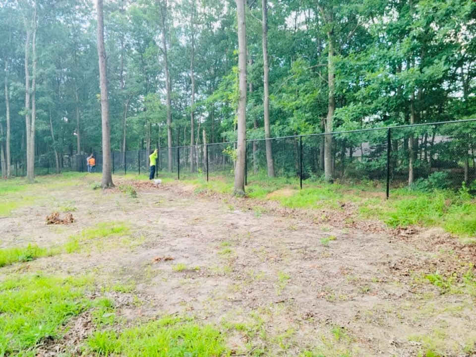 A person wearing safety gear works on a black fence in a forest clearing. Green trees surround the area.