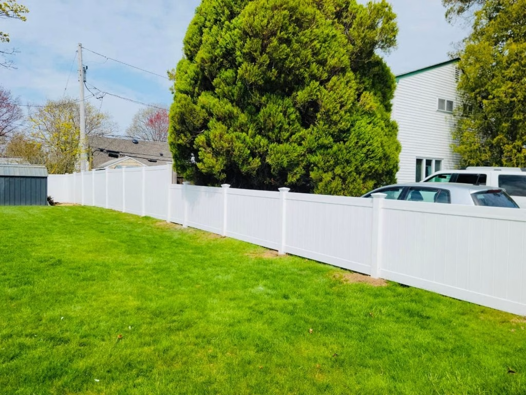 White vinyl fence in a grassy yard, with a large green tree behind it and a white building.