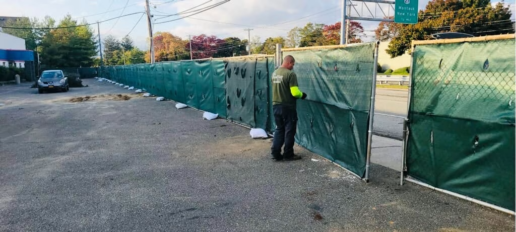 A person next to a green tarp fence. Gravel parking area. Green foliage in the background.