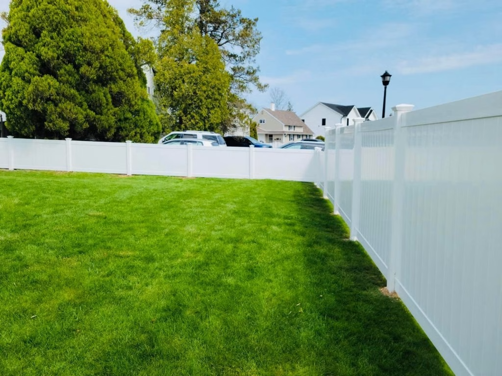 Green lawn bordered by a white fence under a partly cloudy sky; houses and cars in background.