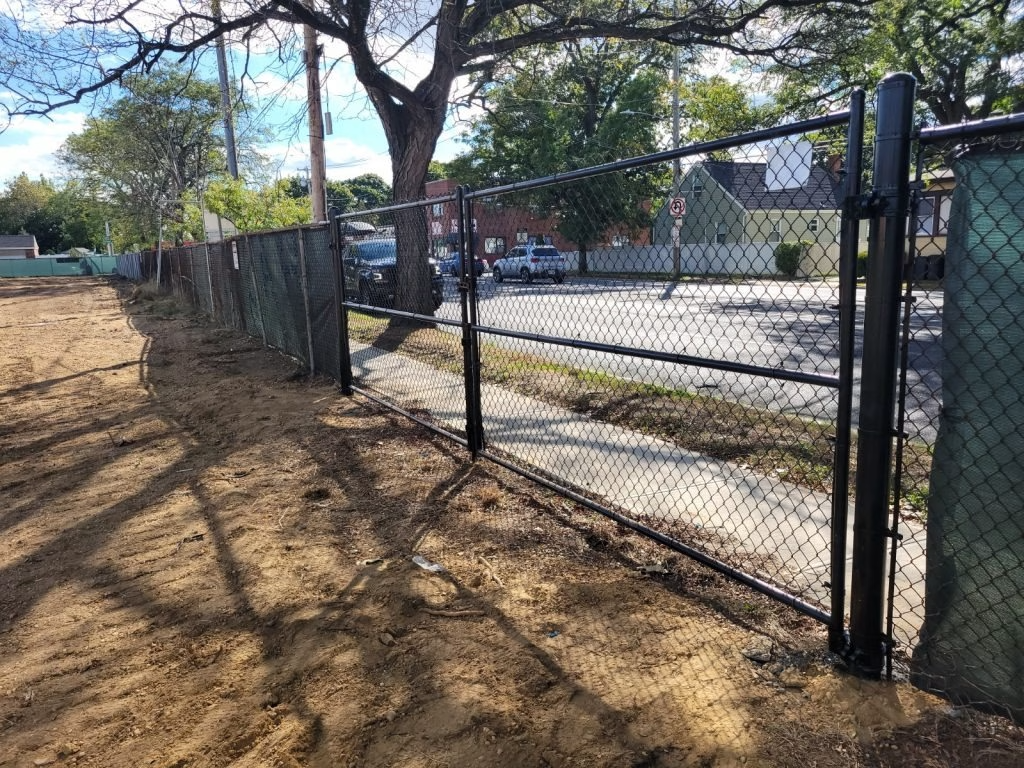 Black chain-link fence with gate, bordering a dirt area. Green foliage visible in the background.