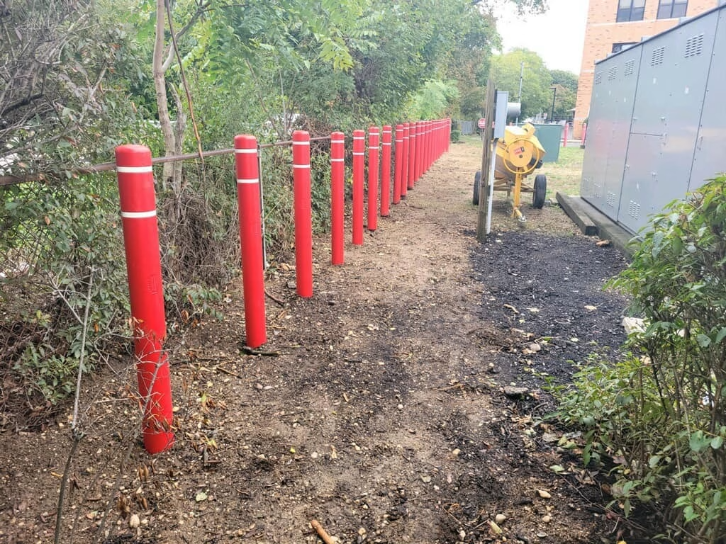 Red bollards with white stripes line a dirt path next to vegetation, near a cement mixer and building.
