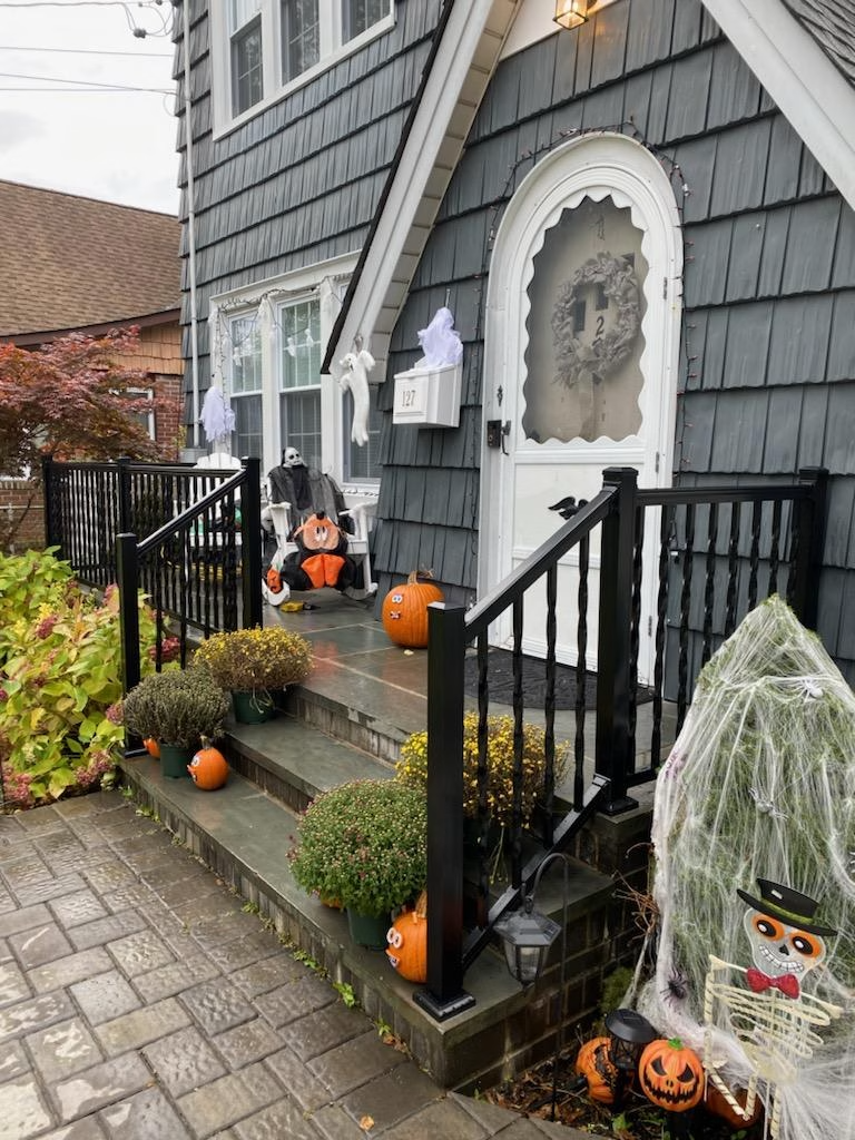 Halloween-decorated house with pumpkins, ghosts, and a scarecrow on the porch and steps. Gray siding and black railing.