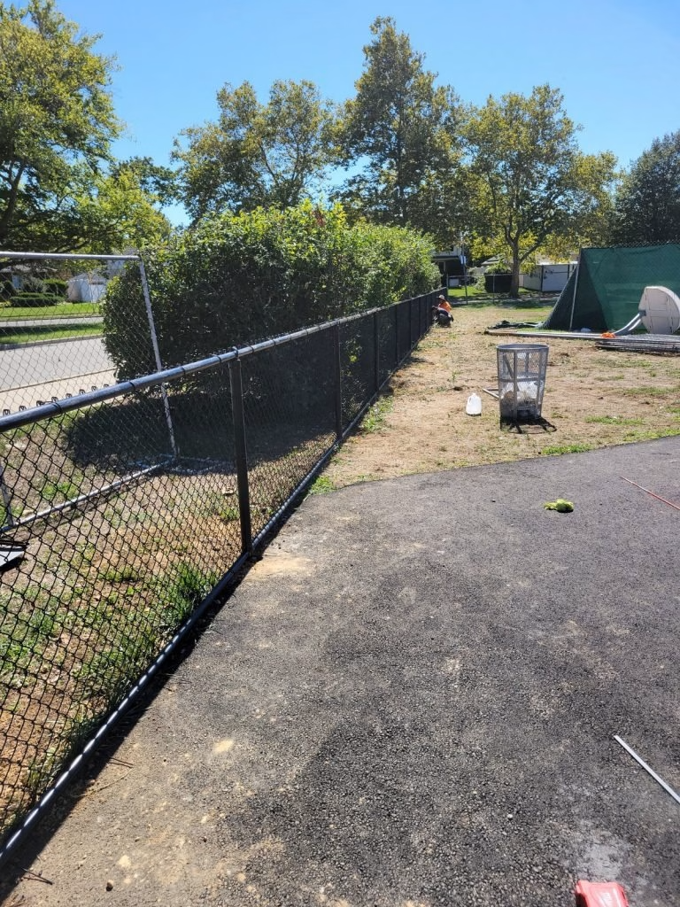 Black chain-link fence bordering a grassy area and paved path, with trees and a cage in the background on a sunny day.