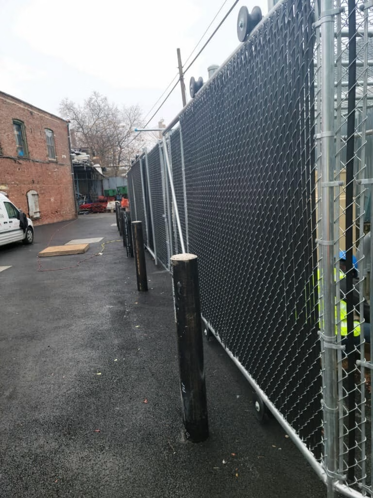 Black chain-link fence with privacy screening, along a paved alleyway with buildings and poles.