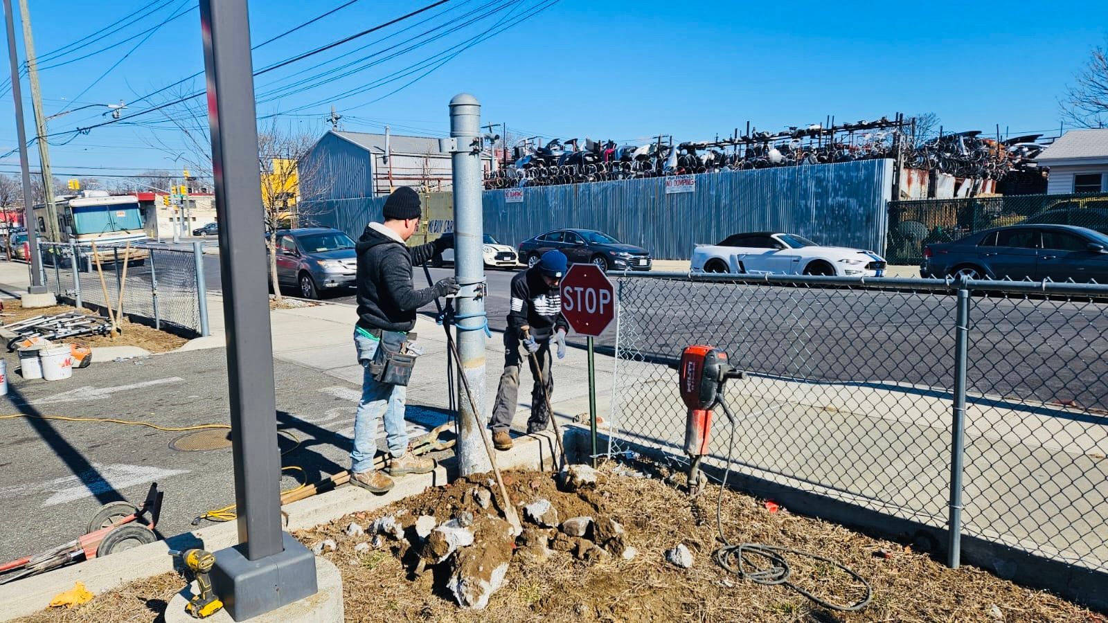 Three construction workers installing a pole next to a stop sign on a sunny day. A junkyard is in the background.