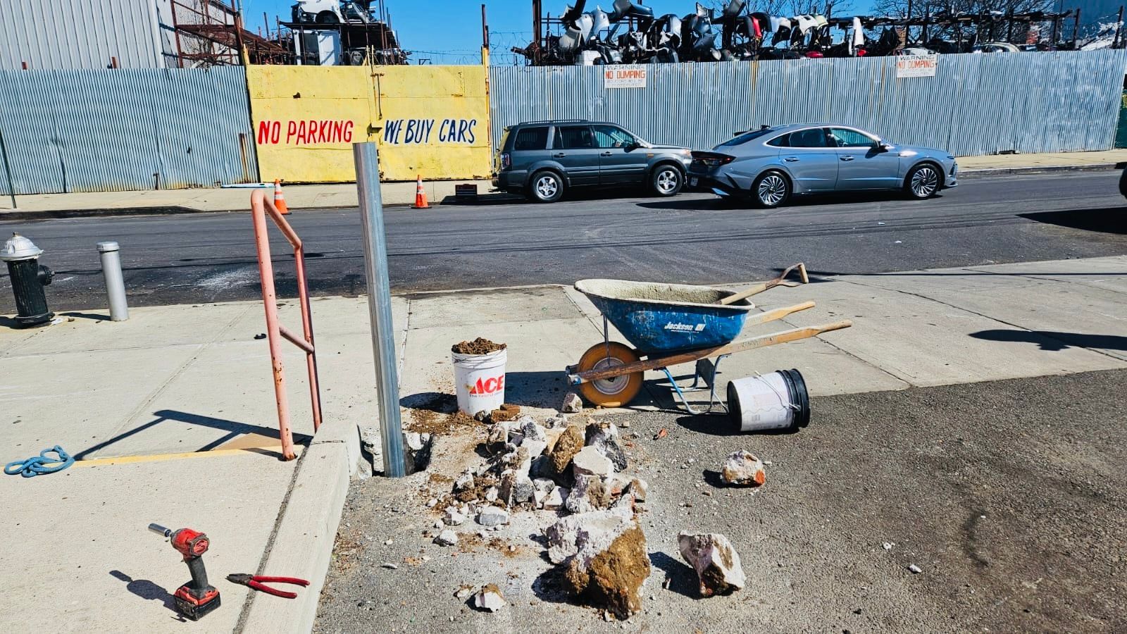 Construction site with tools, debris, and wheelbarrow on a sidewalk. Cars and a fence are in the background.