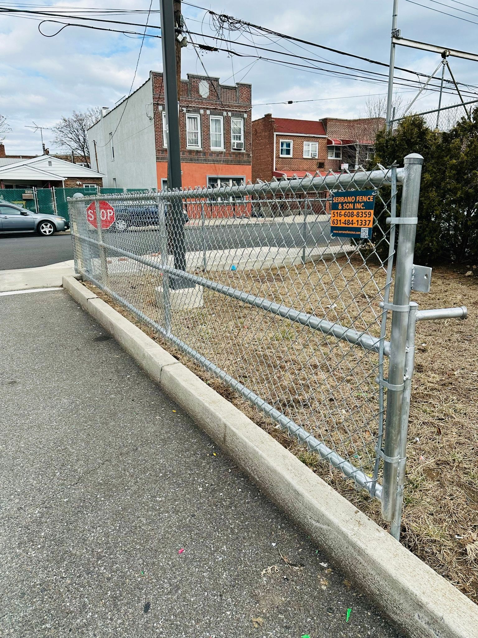 Chain link fence blocking access to a dirt area, with buildings and parked cars in the background.