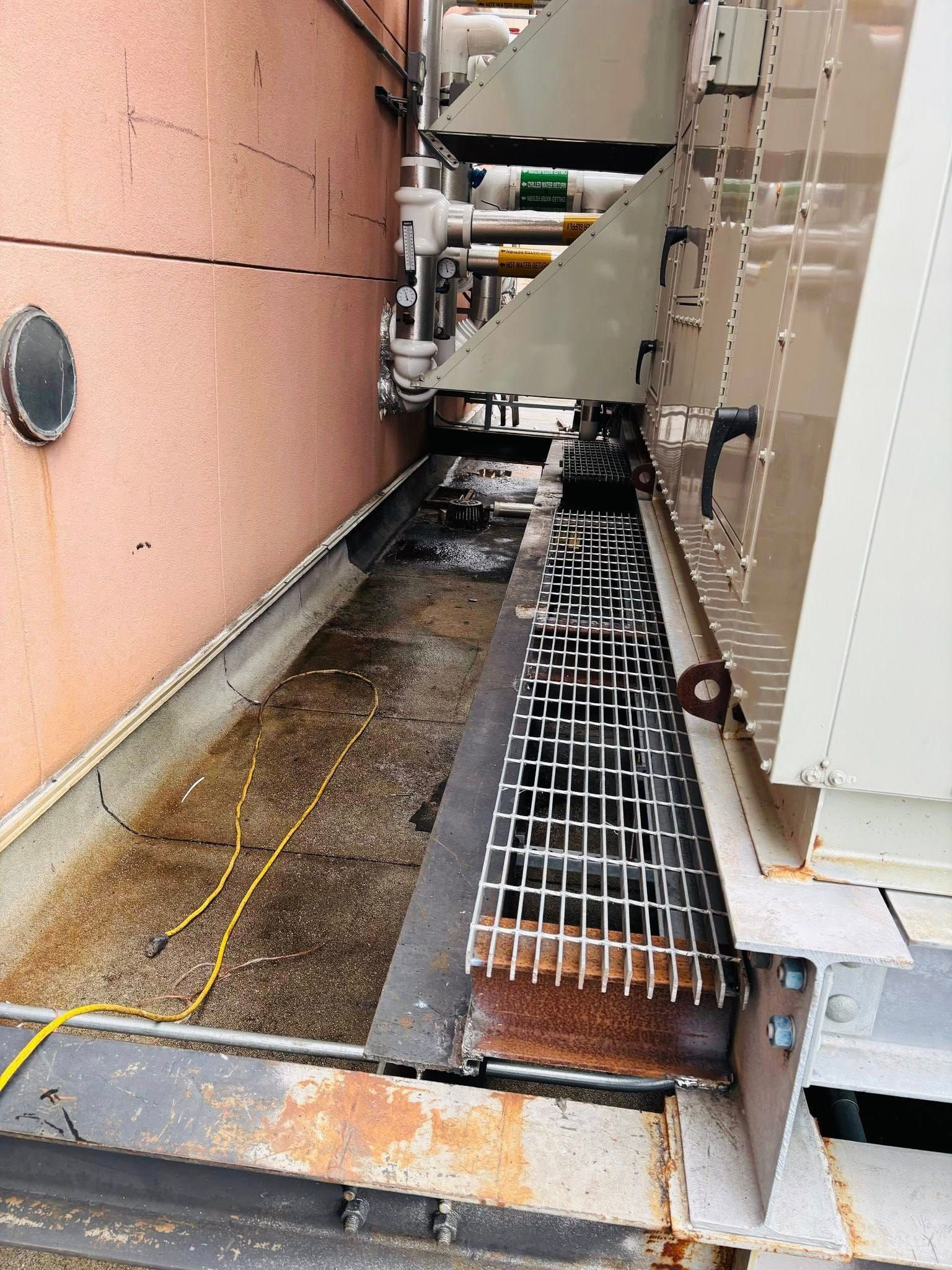 Exterior view of a building's narrow metal walkway next to machinery and a reddish-brown wall with a small round window.