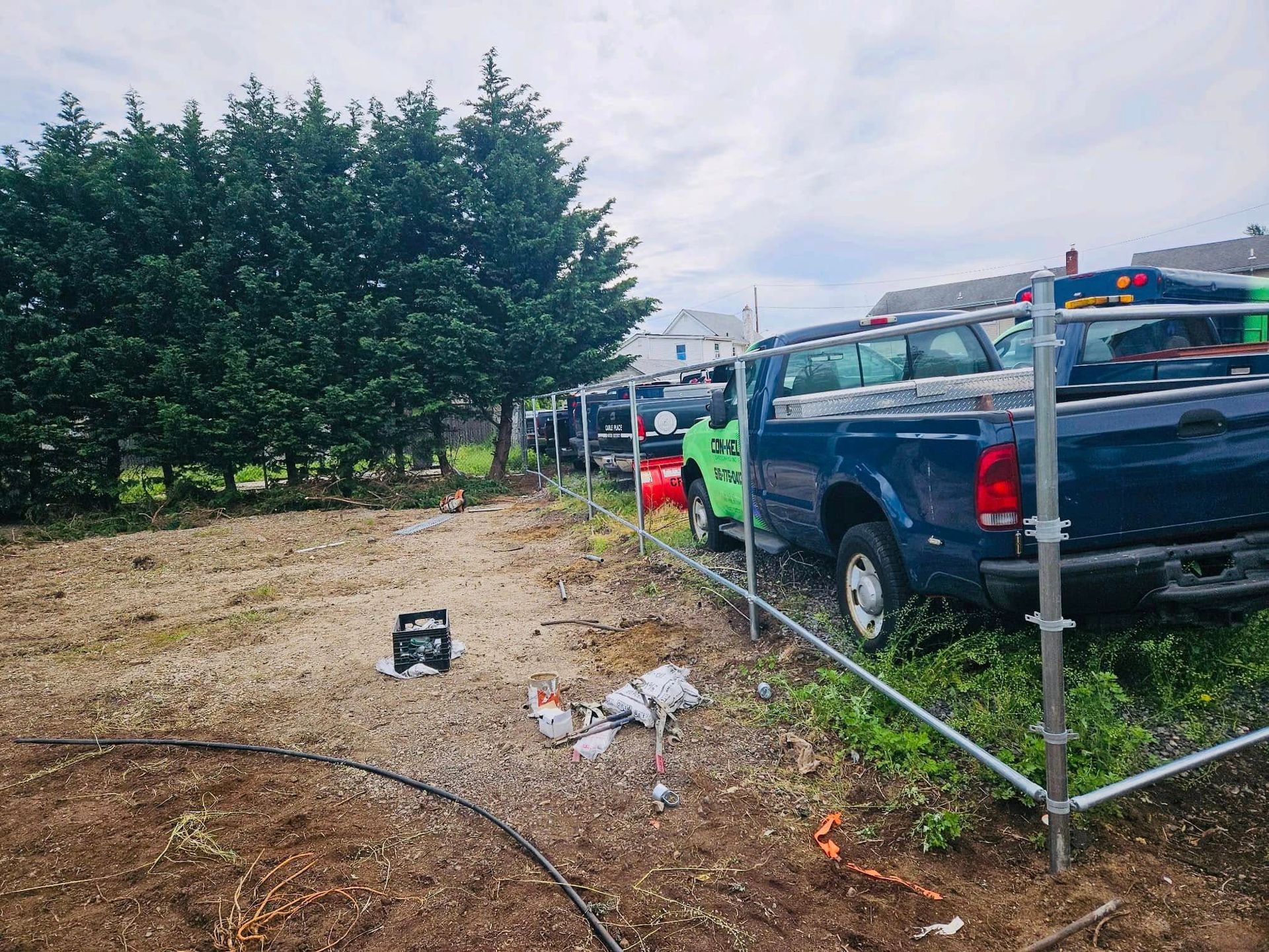 Construction site with trucks, a green van, scaffolding, and a wooded fence line.