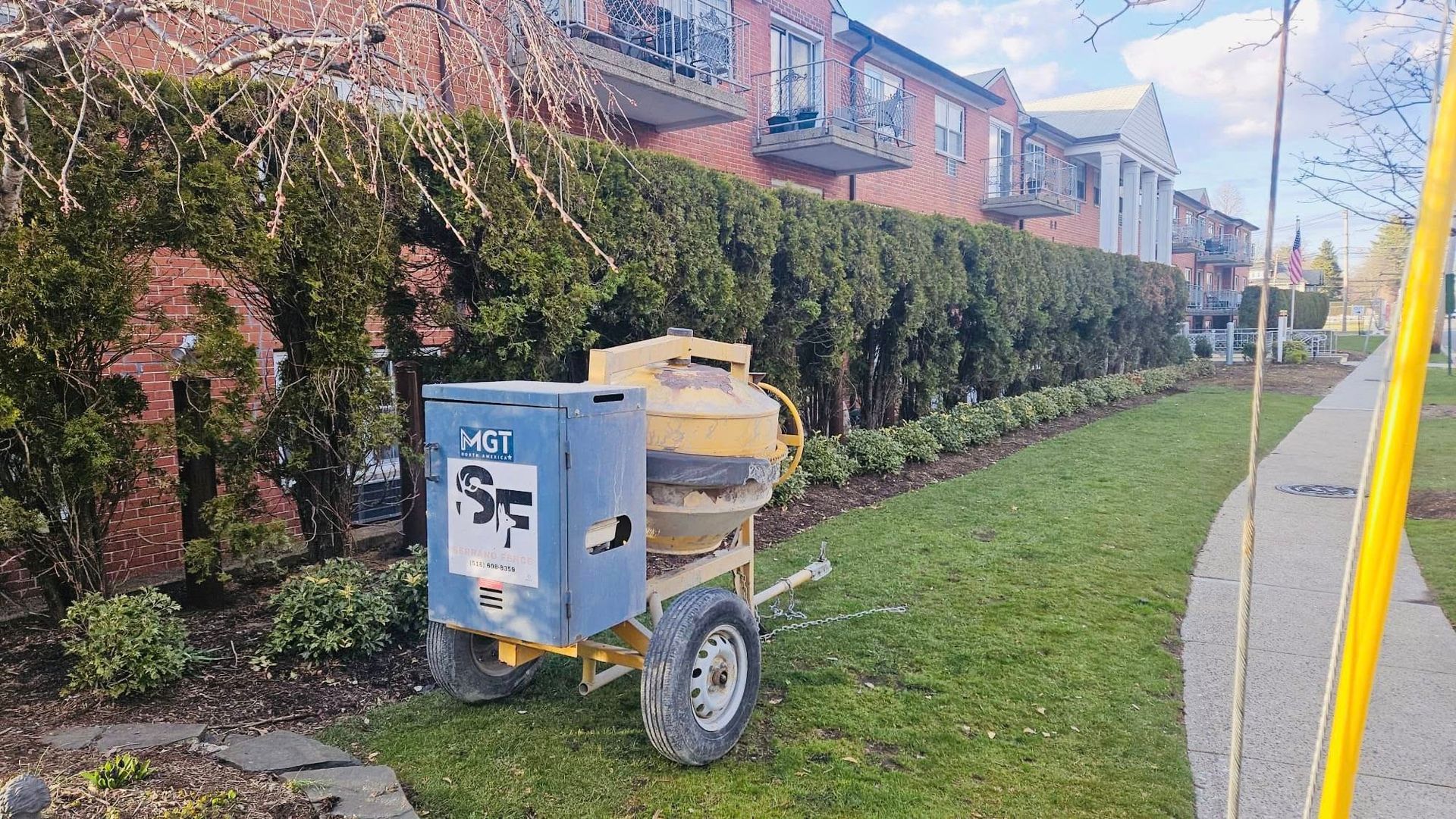 Cement mixer on grass next to a sidewalk and hedge row in front of a brick building.