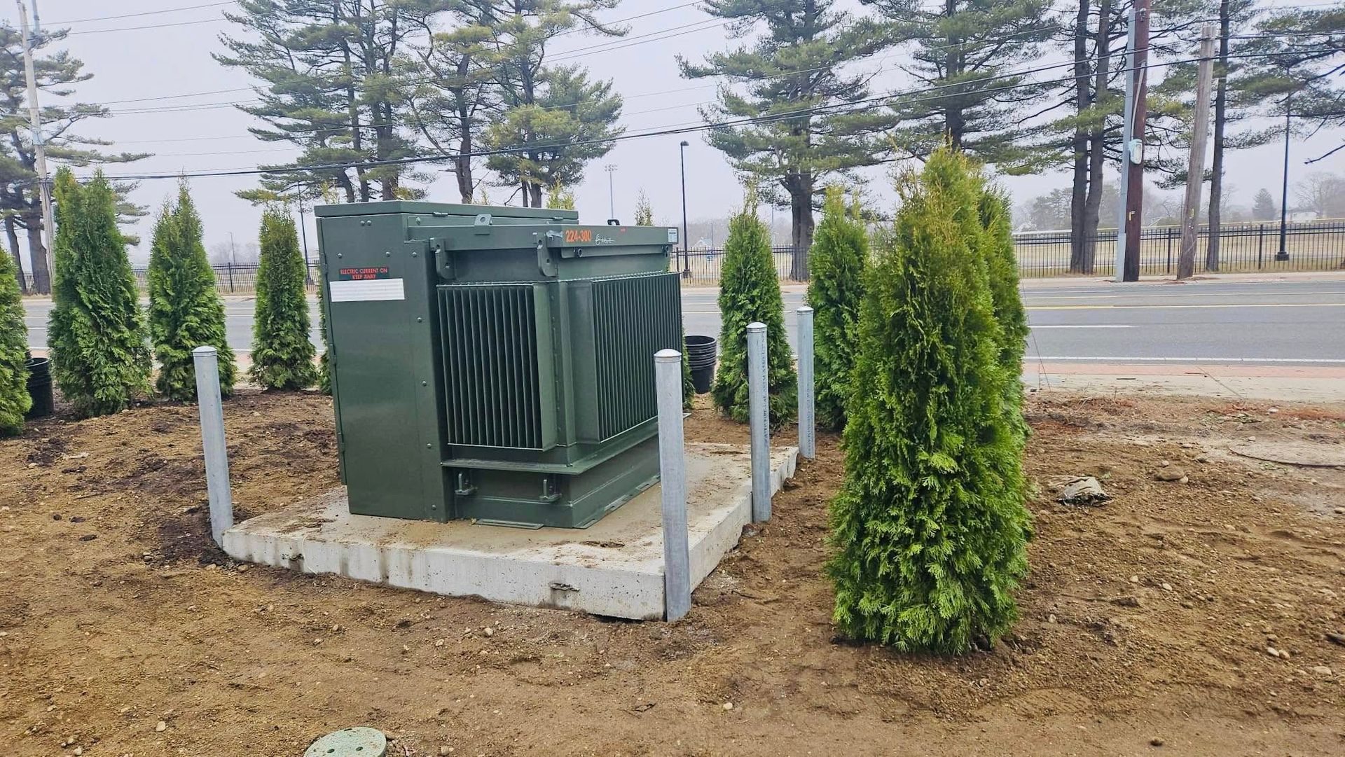 Green electrical transformer box with evergreen trees, metal poles, and roadway in background.