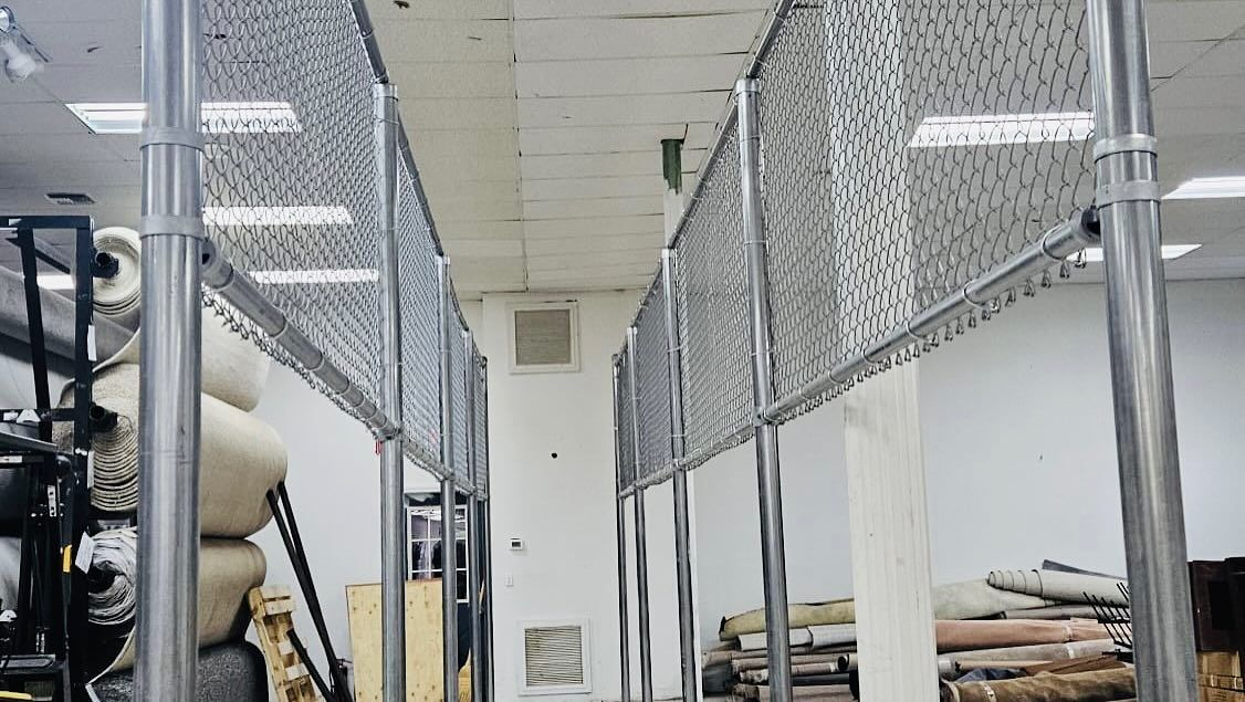 Metal security fencing dividing an interior space, likely a warehouse. Light-colored walls and ceiling.