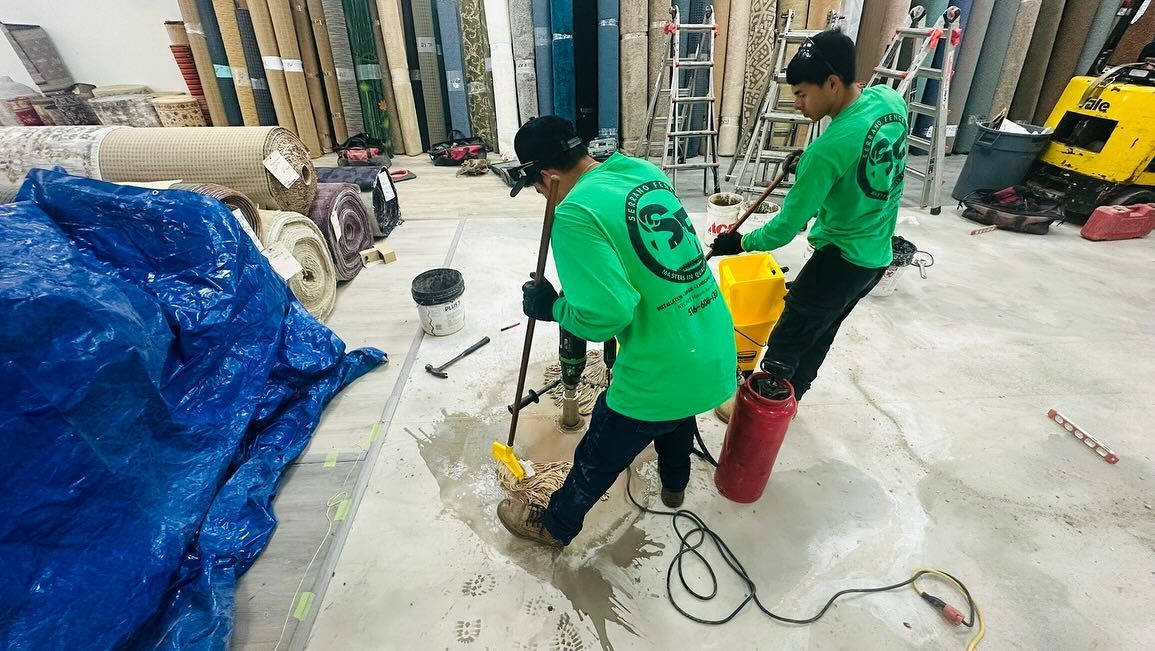 Two workers in green shirts cleaning a flooded warehouse floor with equipment, near a blue tarp and carpet rolls.