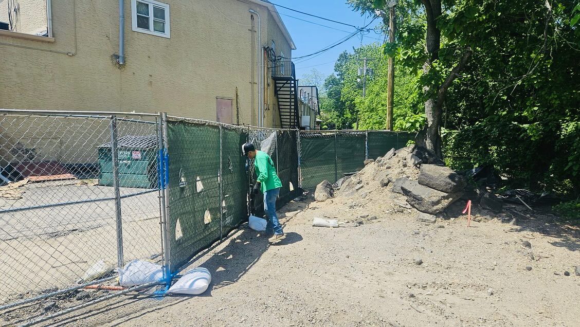 Construction site: a person in green shirt by a chain-link fence, building in background, dirt and gravel ground.