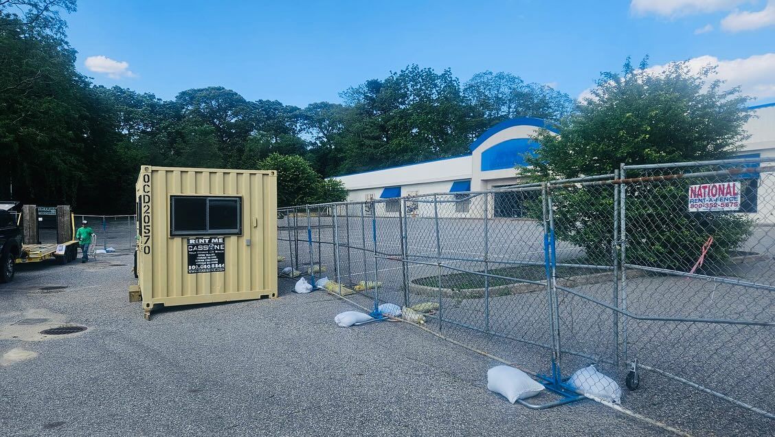 A tan shipping container with a window sits in a gravel lot next to a fenced, closed building under a blue sky.