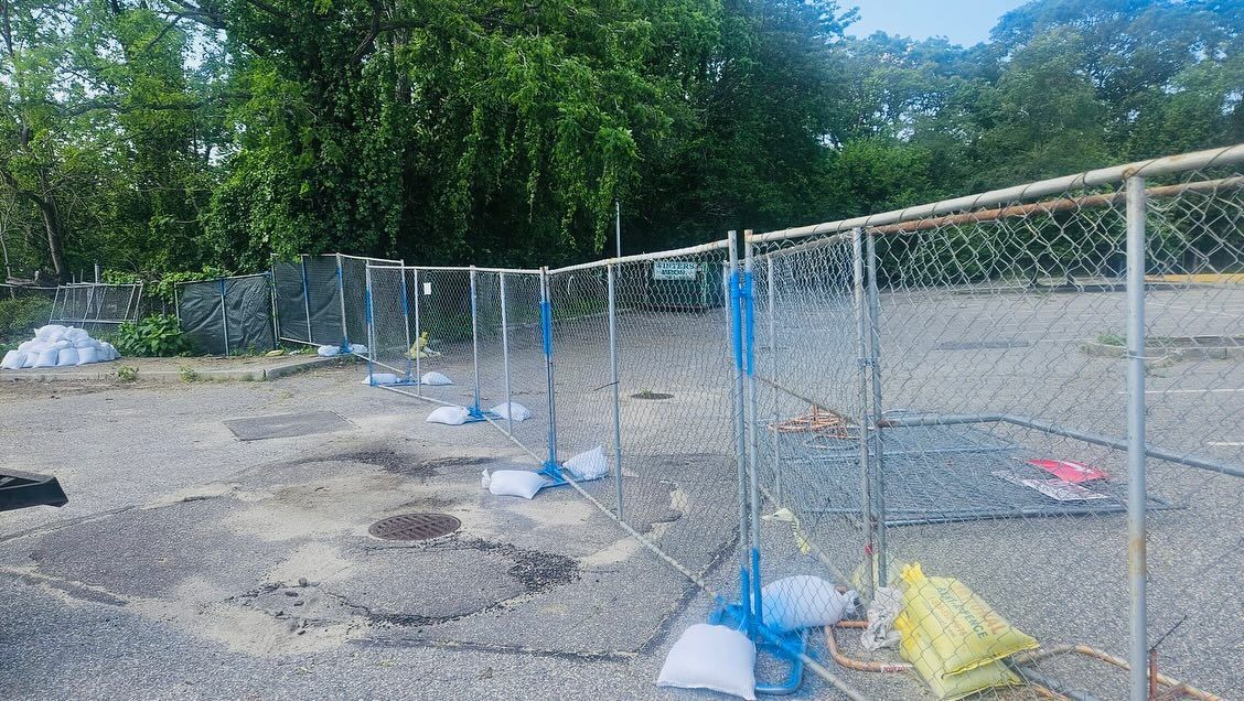 Chain-link fence in a paved lot, with blue and yellow safety barriers attached. Trees in the background.