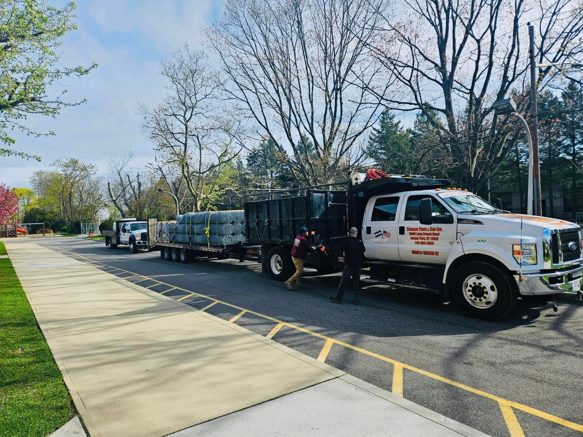 Truck with trailer carrying cargo parked on a street. A person is near the truck. Trees and building are in the background.
