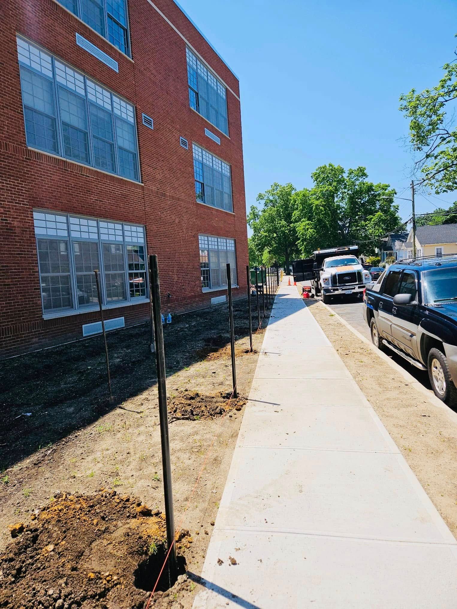 Sidewalk next to brick building, with new trees planted along the edge. A truck and car are on the street.