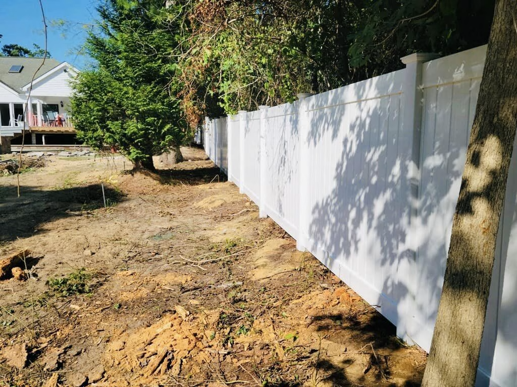 White fence bordering a yard with trees and house in the background. Brown soil in foreground.