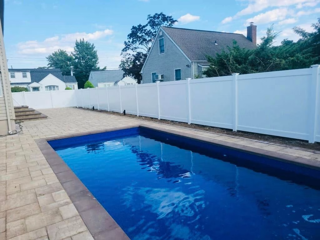 Pool in a fenced backyard, with a brick patio and a house in the background.