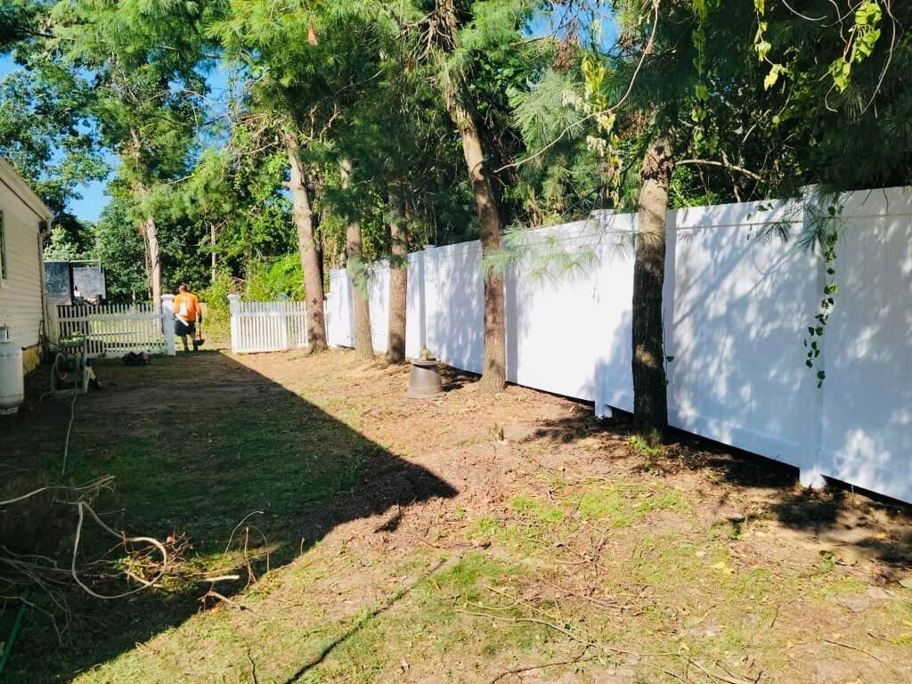 White fence lines a backyard with trees; person in orange shirt works in the distance.