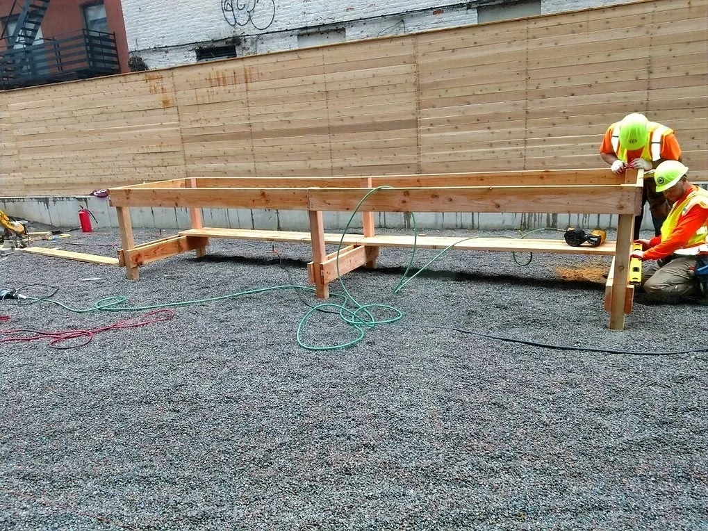 Construction workers on gravel next to a wooden work table. Yellow vests, hard hats, and tools are visible.