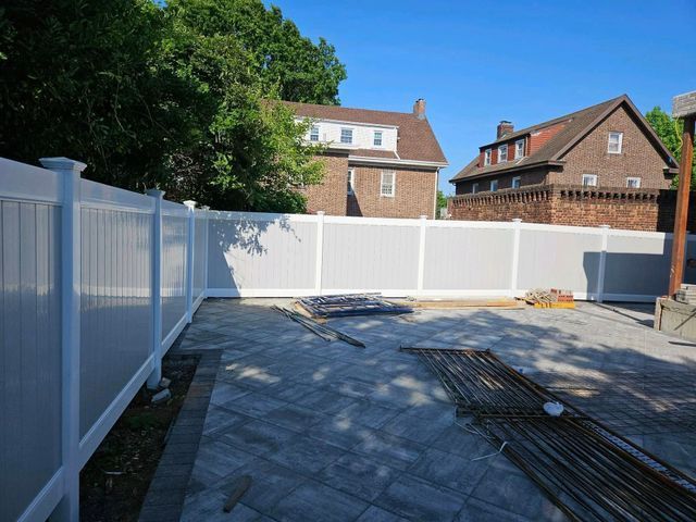 Backyard with white vinyl fence, paved patio, and brick houses under a sunny sky