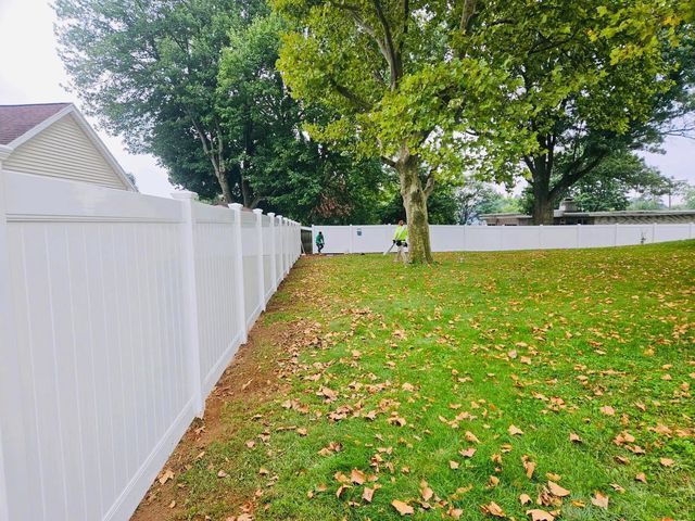 White fence beside a grassy backyard with scattered leaves and a large tree.