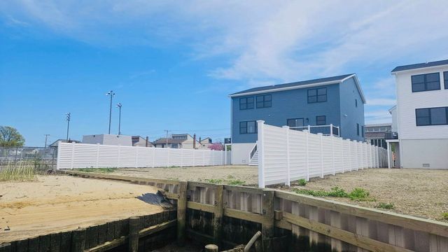 Canal with wooden dock in foreground, sandy lot, and blue houses under clear sky