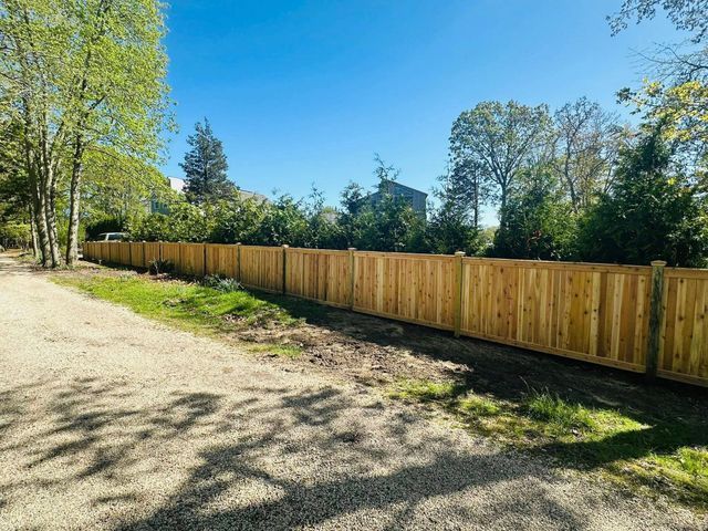 Gravel driveway beside a long wooden fence, with trees and a bright blue sky.