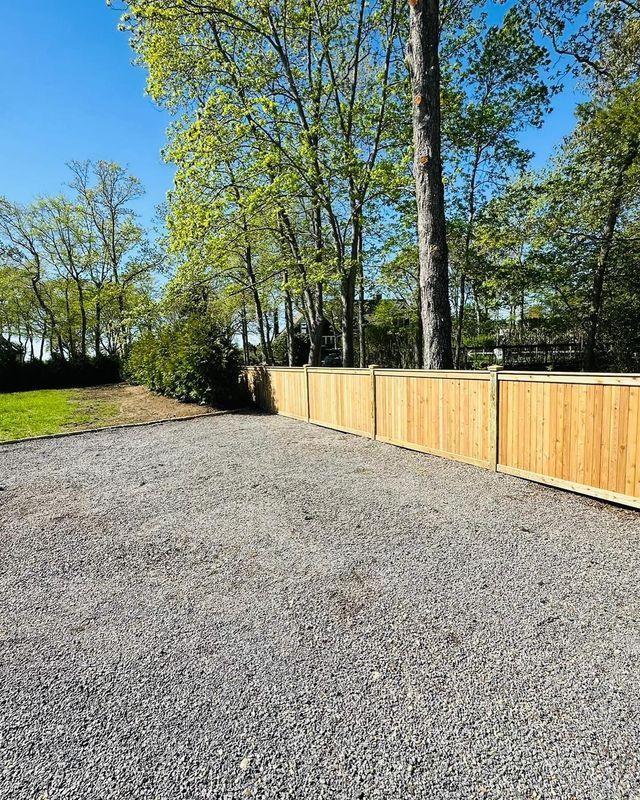 Gravel driveway beside a wooden fence, with trees and blue sky in the background.
