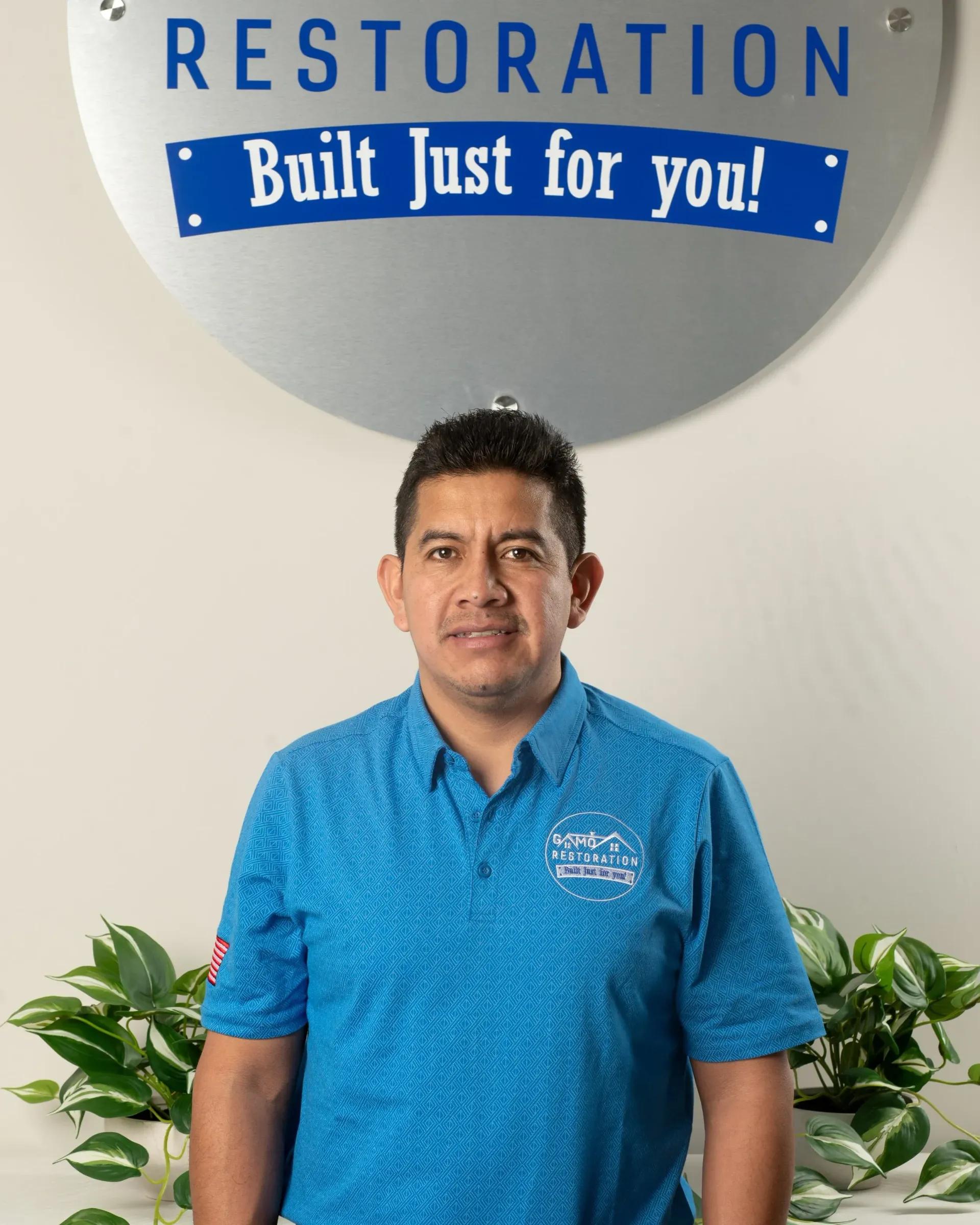 Man standing in blue polo in front of Restoration sign, indoors with plants on either side