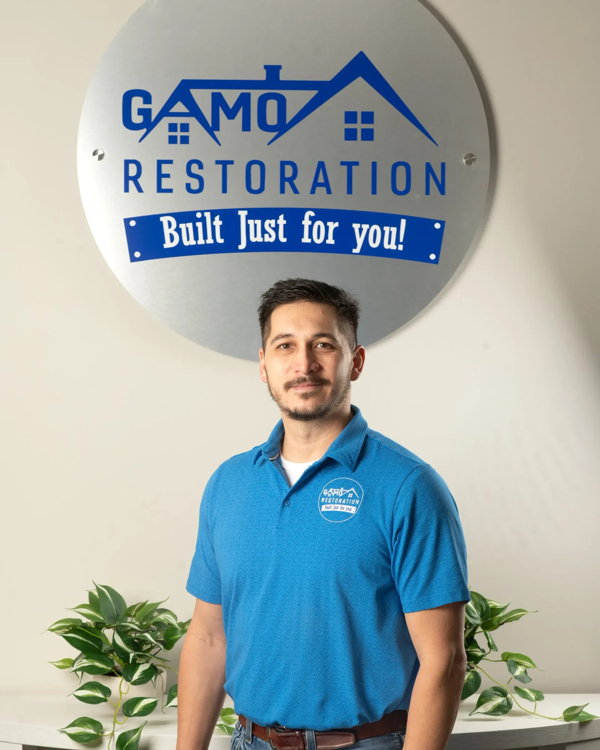 Man standing in front of a GAMO Restoration sign, wearing a blue polo shirt in an office setting