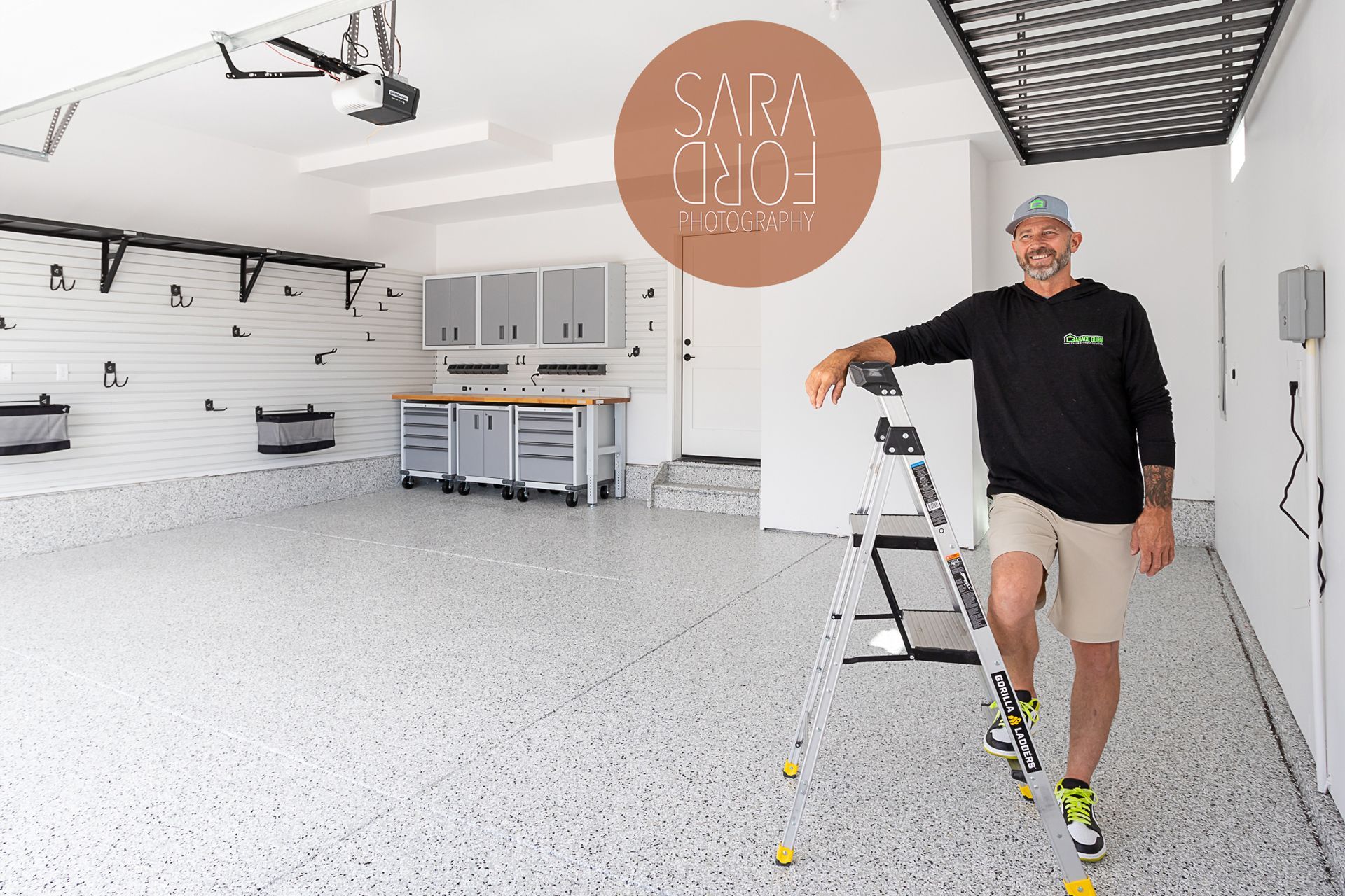 Man in garage with epoxy floor, leaning on ladder. Beige shorts, green shoes. Brown cabinetry in background.