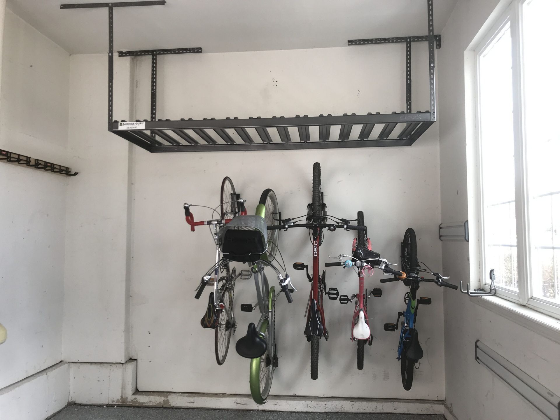 Garage with bikes hanging on wall, storage shelf above. White walls, window to the right.