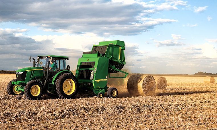 Green tractor pulling a hay baler, creating round bales in a field. Sunny day.