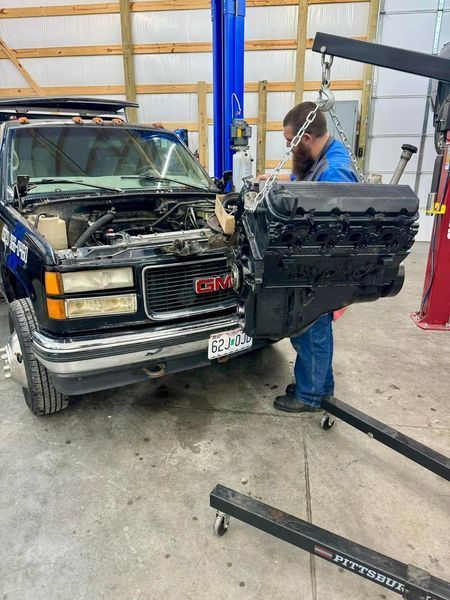 Mechanic removing a black engine from a GMC truck with a hoist in a garage.