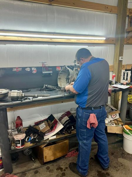 Mechanic works on car part at a workbench in a shop. He wears blue shirt, jeans.