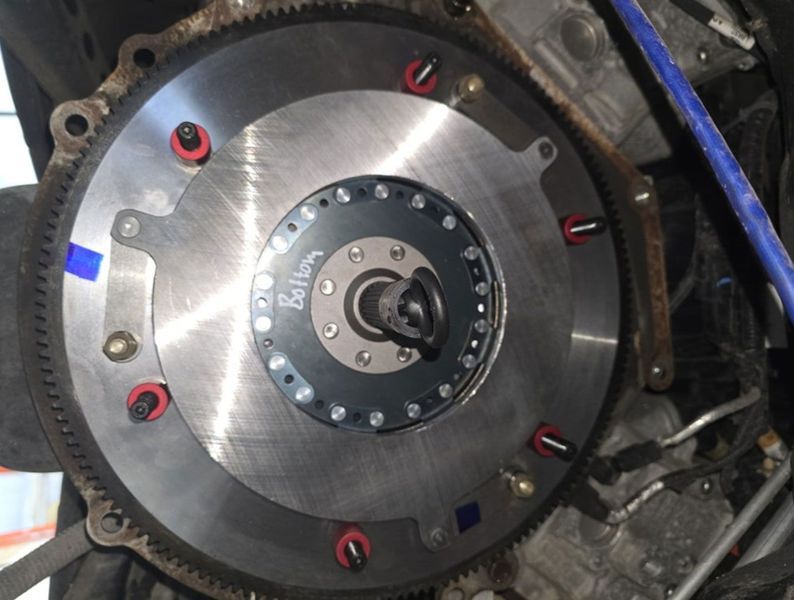 A silver flywheel with a black clutch assembly, bolts visible, likely in an engine bay.