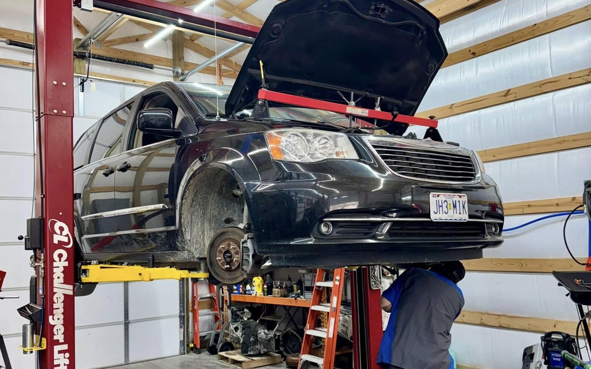 Black minivan on a lift in a garage with the hood open, a person is working underneath.
