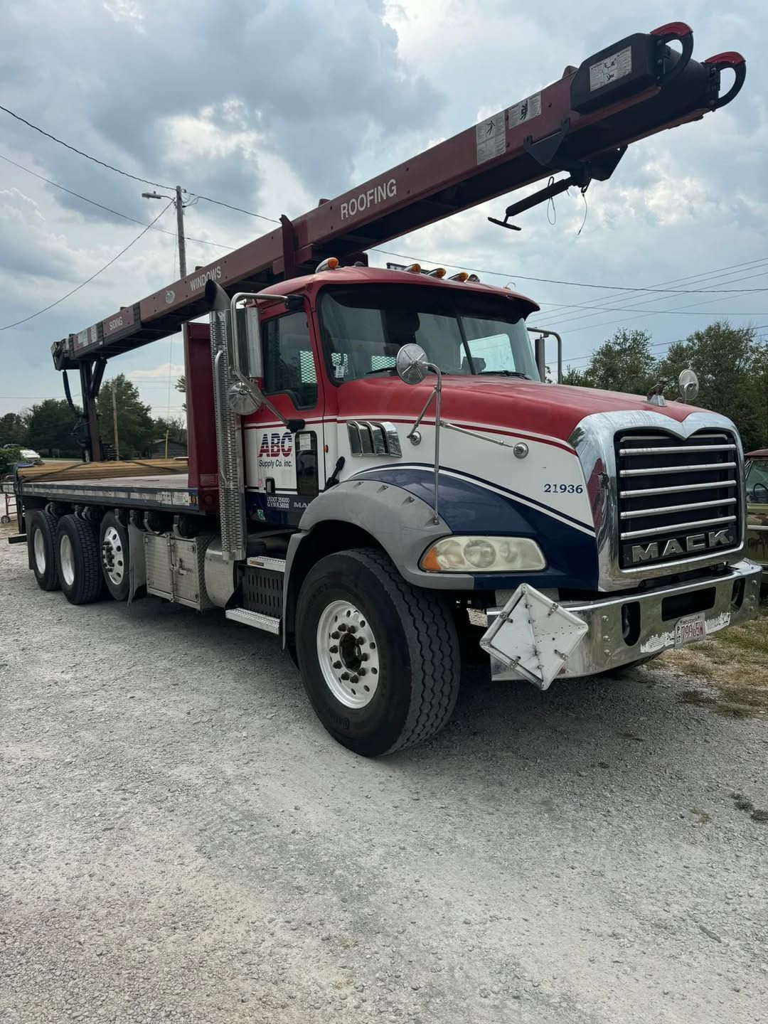 Red, white, and blue Mack truck with a crane boom, parked on gravel.
