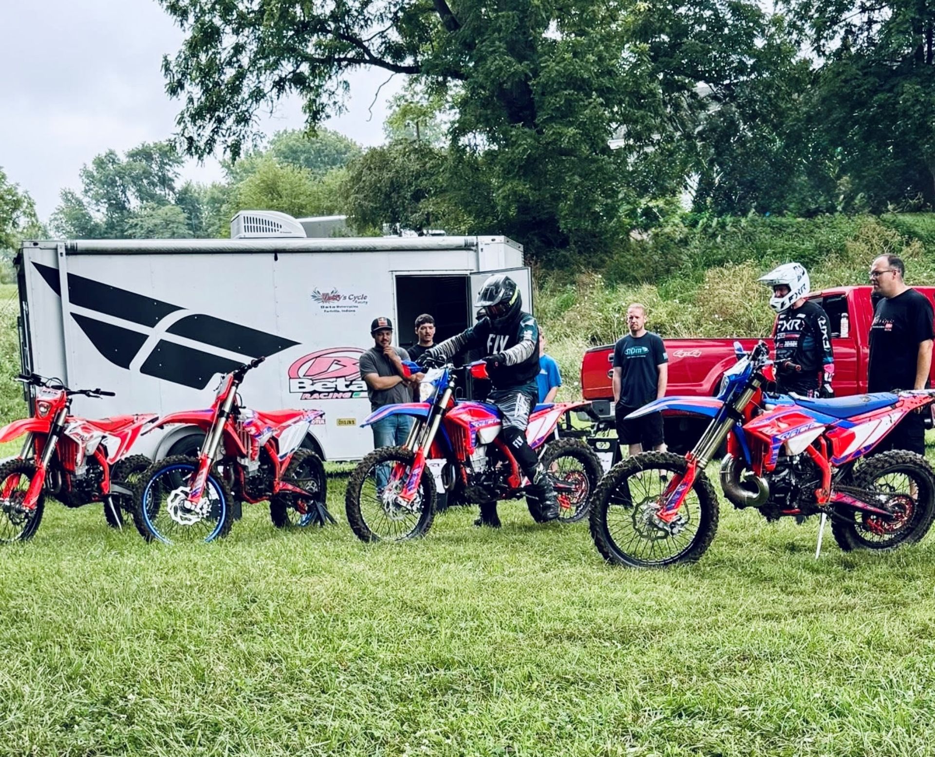 Children on dirt bikes lined up on grass near a trailer, with riders and a red pickup in the background