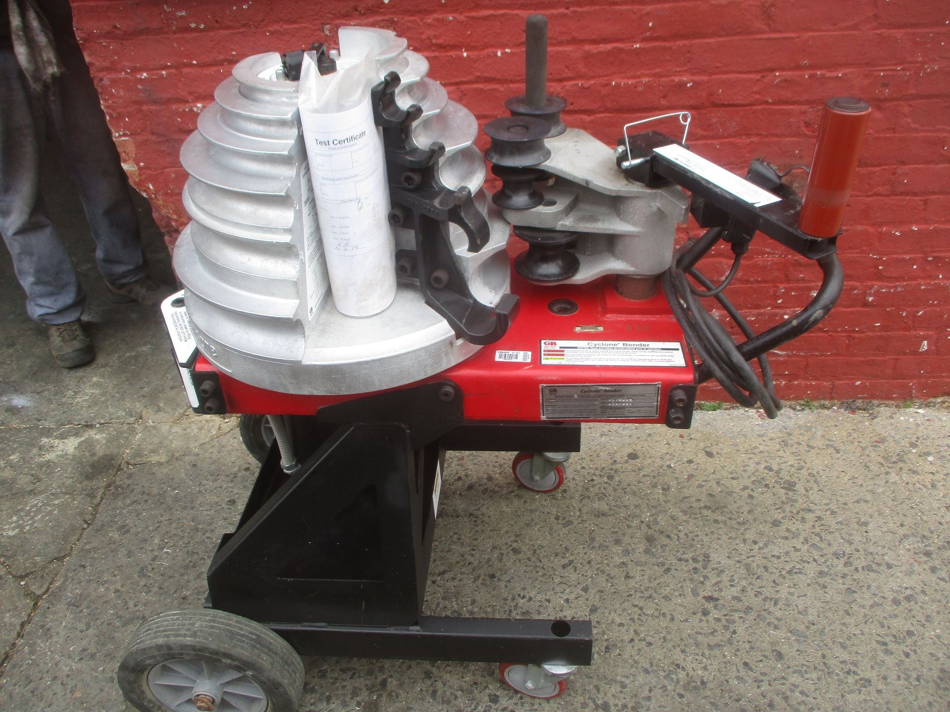 A red and black pipe bending machine is sitting in front of a red brick wall