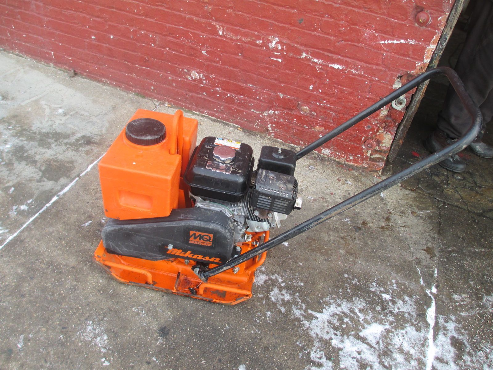 A small orange machine is sitting on the ground in front of a red brick wall.