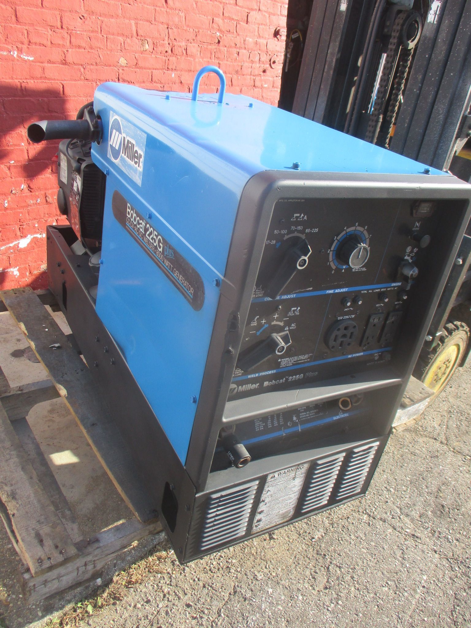 A blue welding machine is sitting on a pallet in front of a red brick wall.